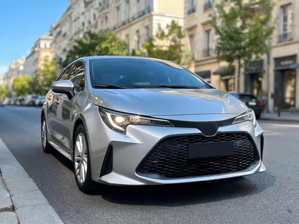Srebrny kompakt klasy Toyota Corolla zaparkowany na słonecznej, miejskiej ulicy, podkreślający przód auta i reflektory.